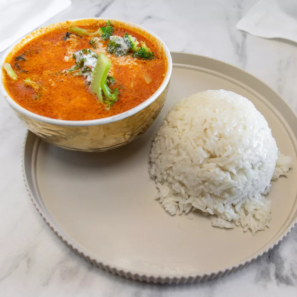 A bowl of red Thai curry with broccoli and a side of white rice on a light plate.