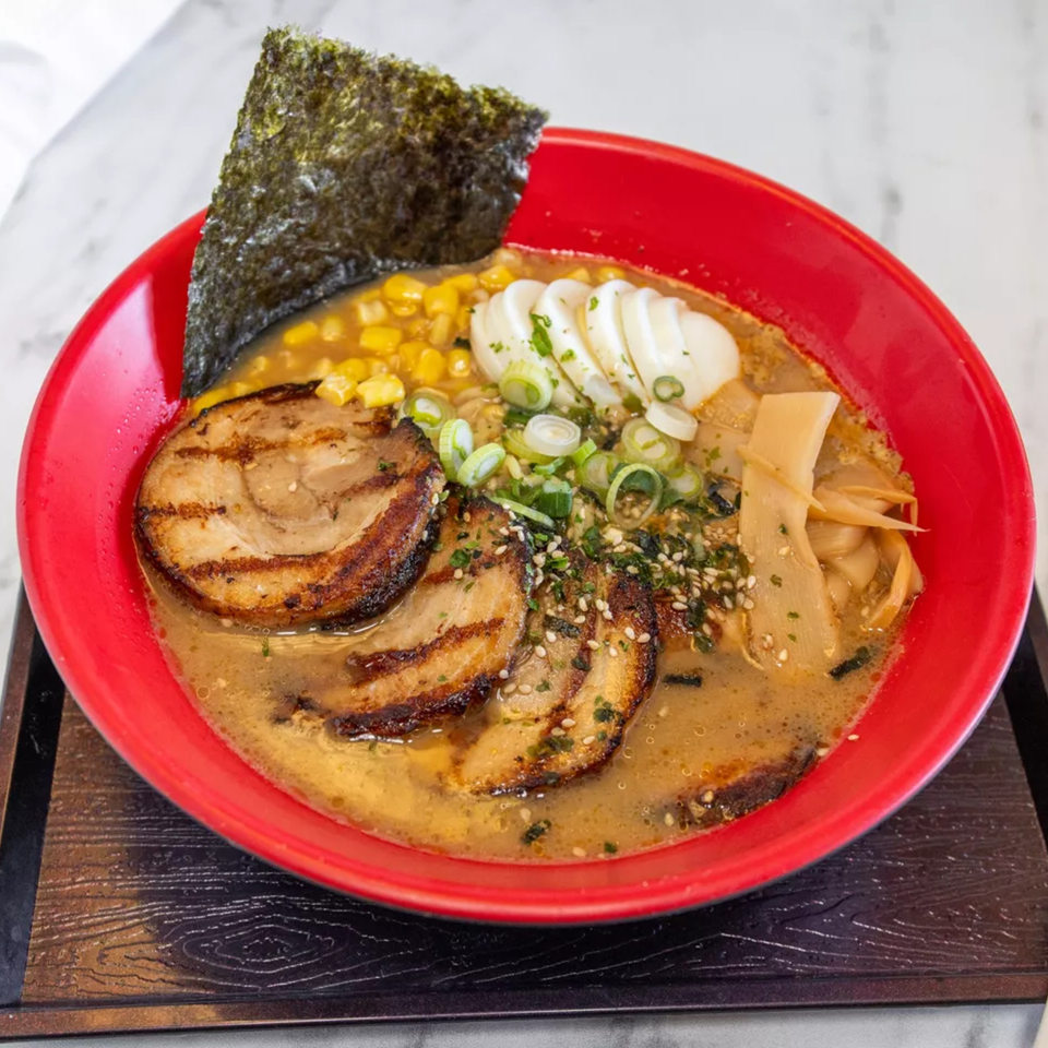 Ramen in a red bowl with pork, corn, egg, green onions, bamboo shoots, and nori.
