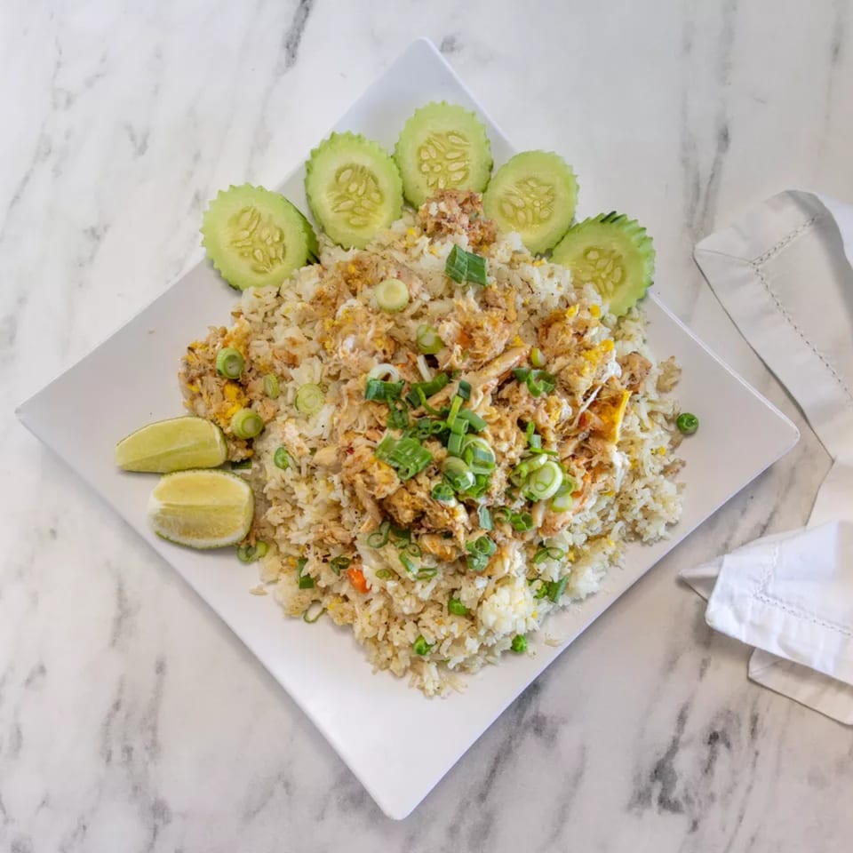 Crab fried rice with sliced cucumbers, lime wedges, and green onions on a white plate.